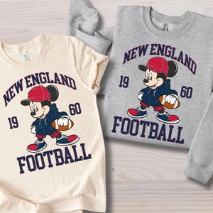 May include: Two t-shirts featuring a cartoon Mickey Mouse in a football uniform. The shirts are cream and gray, with the text "NEW ENGLAND FOOTBALL 1960". Mickey is wearing a red cap, blue hoodie, and holding a football.