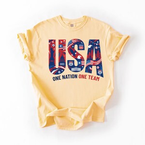 May include: A pale yellow short-sleeve t-shirt featuring a large graphic with the letters USA in blue and red. The letters contain illustrations of the Statue of Liberty, the White House, eagles, and Route 66 signs. Below is the text ONE NATION ONE TEAM.