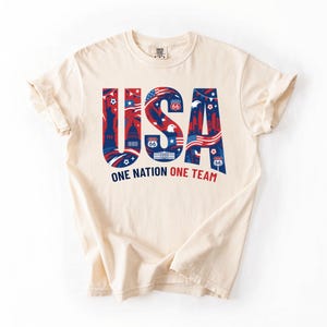May include: A cream-colored short-sleeve t-shirt featuring the large blue and red text USA. The letters contain graphic illustrations of the Statue of Liberty, a bald eagle, Route 66 signs, and the American flag. Below is the text ONE NATION ONE TEAM.