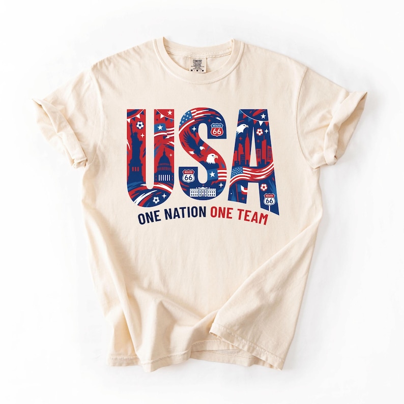May include: A cream-colored short-sleeve t-shirt featuring the large blue and red text USA. The letters contain graphic illustrations of the Statue of Liberty, a bald eagle, Route 66 signs, and the American flag. Below is the text ONE NATION ONE TEAM.
