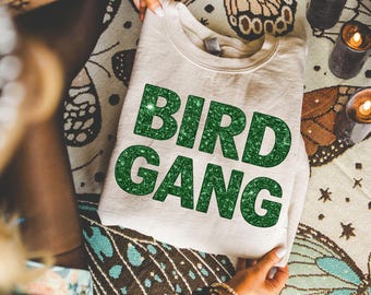 May include: A beige crewneck sweatshirt featuring the text BIRD GANG printed in large, bold, glittery green letters across the chest. The garment has a classic long-sleeve design with a ribbed collar, cuffs, and hem, made of a soft fabric.