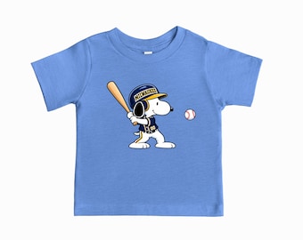 Puede incluir: Una camiseta de manga corta azul claro con un gráfico de un perro de dibujos animados que lleva un uniforme y casco de béisbol azul y amarillo con la palabra MILWAUKEE, sosteniendo un bate de madera y preparándose para golpear una pelota.