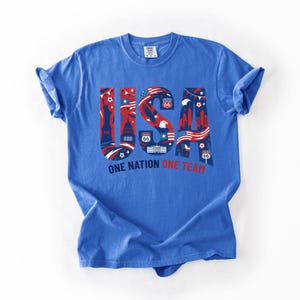May include: A blue short-sleeve t-shirt featuring a large graphic with the letters USA in red, white, and blue. The design includes the Statue of Liberty, the White House, bald eagles, and Route 66 signs. Below the graphic is the text ONE NATION ONE TEAM.