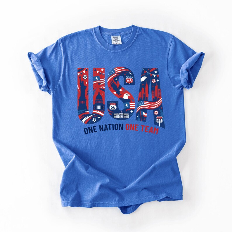 May include: A blue short-sleeve t-shirt featuring a large graphic with the letters USA in red, white, and blue. The design includes the Statue of Liberty, the White House, bald eagles, and Route 66 signs. Below the graphic is the text ONE NATION ONE TEAM.