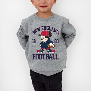 May include: Gray sweatshirt featuring a cartoon Mickey Mouse in a football uniform. The design includes the words "NEW ENGLAND FOOTBALL" and the numbers "19" and "60". The sweatshirt has a crew neck and long sleeves.