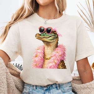 Puede incluir: Camiseta color crema con una ilustraci&oacute;n detallada de un caim&aacute;n con gafas de sol rosas, una boa rosa y una cadena dorada. El caim&aacute;n tiene una piel texturizada verde y marr&oacute;n. La camiseta es una prenda casual.