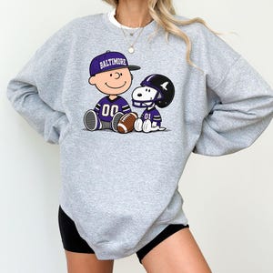May include: A heather gray crewneck sweatshirt featuring a graphic of a cartoon character wearing a purple Baltimore baseball cap and a purple jersey with the number 00, sitting next to a cartoon dog wearing a purple football helmet and jersey.