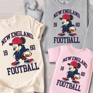 May include: Several t-shirts and a sweatshirt featuring a cartoon Mickey Mouse in a football uniform. The shirts are cream, gray, and pink, with the words "New England Football" and the numbers "19" and "60".
