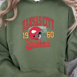 K&ouml;nnte beinhalten: Gr&uuml;ner Kapuzenpullover mit einem Vintage-Grafikdesign mit dem Text "Kansas City Football" mit einem roten Footballhelm und den Zahlen 19 und 60.