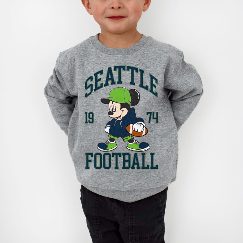May include: A gray sweatshirt featuring a graphic of Mickey Mouse in a football uniform. The design includes the text "SEATTLE FOOTBALL" and the numbers "19" and "74". The sweatshirt is worn by a child.