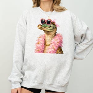 Puede incluir: Una sudadera gris claro con una ilustraci&oacute;n caprichosa de un caim&aacute;n con gafas de sol rosas y una boa rosa. El caim&aacute;n tiene un collar de cadena dorada. La sudadera es de estilo c&oacute;modo e informal.