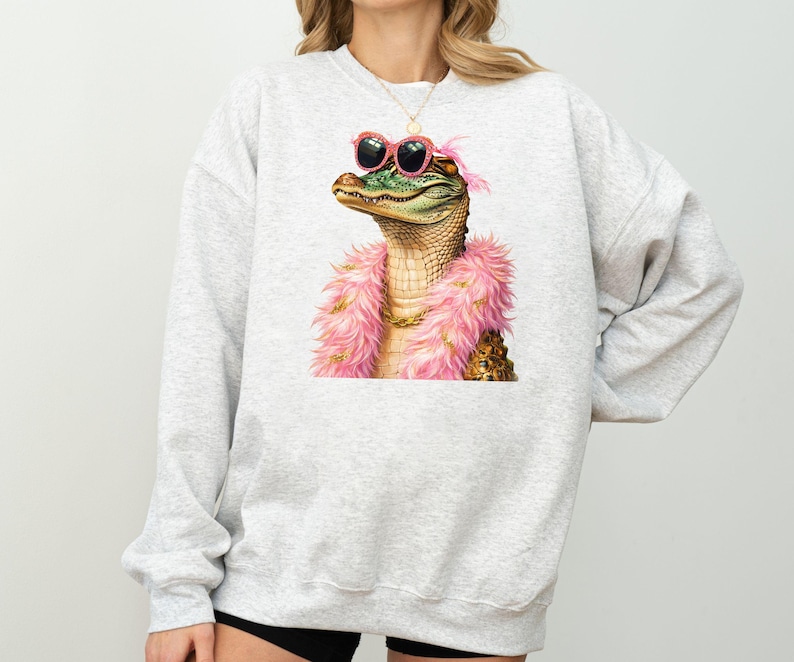 Puede incluir: Una sudadera gris claro con una ilustraci&oacute;n caprichosa de un caim&aacute;n con gafas de sol rosas y una boa rosa. El caim&aacute;n tiene un collar de cadena dorada. La sudadera es de estilo c&oacute;modo e informal.