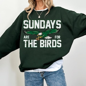 May include: A dark green crewneck sweatshirt featuring a white graphic print that reads SUNDAYS ARE FOR THE BIRDS. The design includes a central illustration of a green and white eagle clutching a brown football in its talons.