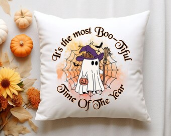 May include: White square pillow with a Halloween-themed design. The design features a cartoon ghost wearing a witch hat and holding a broom and a jack-o'-lantern. Text reads "It's the most Boo-tiful Time Of The Year."