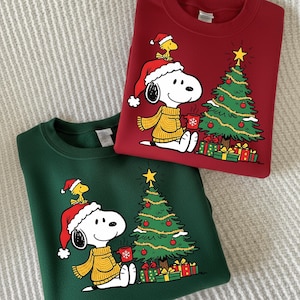 May include: A dark red sweatshirt and a dark green sweatshirt, each featuring a graphic print of Snoopy wearing a Santa hat and yellow sweater, holding a red mug, with Woodstock on their head, a decorated Christmas tree, and gift boxes.