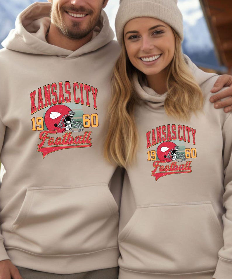 K&ouml;nnte beinhalten: Zwei Personen tragen beigefarbene Hoodies mit einem Vintage-Grafikdruck von Kansas City Football. Die Grafik zeigt einen Footballhelm und den Text "Kansas City 19 60 Football".