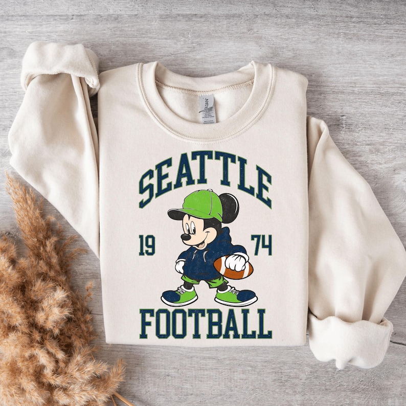 May include: Cream-colored sweatshirt featuring a graphic of Mickey Mouse in a green cap and blue hoodie, holding a football. The text "SEATTLE FOOTBALL 1974" is arched above and below the character in navy blue.