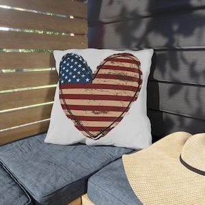 May include: White square pillow with a heart-shaped design featuring the American flag. The heart has a distressed look with red and white stripes and a blue section with white stars. A black outline frames the heart.