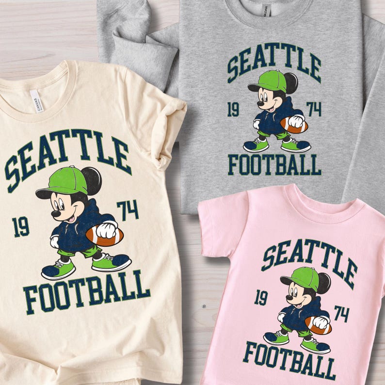 May include: Several t-shirts and a sweatshirt featuring a cartoon character in a football uniform. The shirts are in cream, gray, and pink, with the words "SEATTLE FOOTBALL" and the numbers "19" and "74" in dark green.