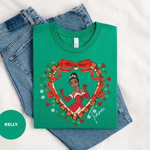 A green t-shirt with a cartoon image of a girl wearing a red dress and a crown, surrounded by a heart-shaped frame with red ribbon and snowflakes. The t-shirt is placed on top of a pair of blue jeans.