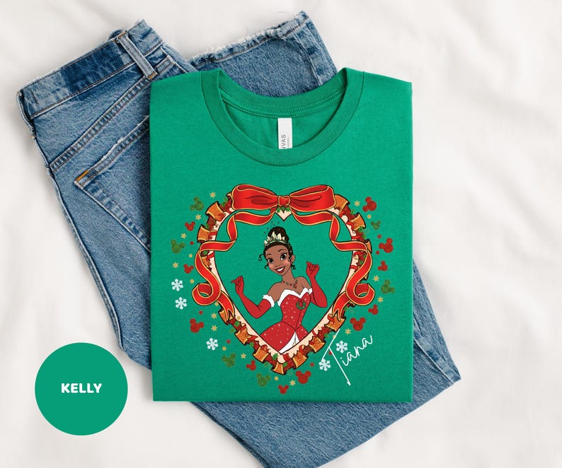 A green t-shirt with a cartoon image of a girl wearing a red dress and a crown, surrounded by a heart-shaped frame with red ribbon and snowflakes. The t-shirt is placed on top of a pair of blue jeans.