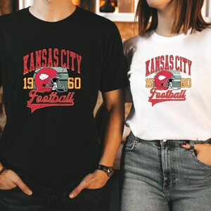 K&ouml;nnte beinhalten: Zwei Personen tragen schwarze und wei&szlig;e T-Shirts mit einem Vintage-Logo des Kansas City Football. Das Logo zeigt einen roten Footballhelm mit einem wei&szlig;en Streifen und dem Text "Kansas City 1960 Football".