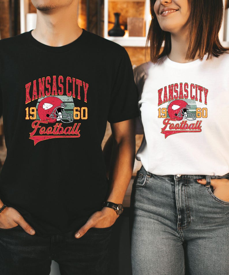 K&ouml;nnte beinhalten: Zwei Personen tragen schwarze und wei&szlig;e T-Shirts mit einem Vintage-Logo des Kansas City Football. Das Logo zeigt einen roten Footballhelm mit einem wei&szlig;en Streifen und dem Text "Kansas City 1960 Football".