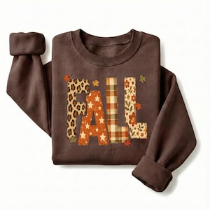 May include: A dark brown long-sleeve sweatshirt featuring the word FALL printed in large block letters. Each letter has a distinct pattern: leopard print, orange with white stars, brown and cream plaid, and a floral design with small autumn leaves.