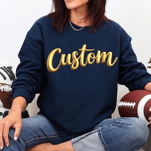 May include: A navy blue crewneck sweatshirt featuring the word Custom printed in a large, three-dimensional gold script font across the chest. The garment has long sleeves and a ribbed neckline, providing a classic fit for casual wear.