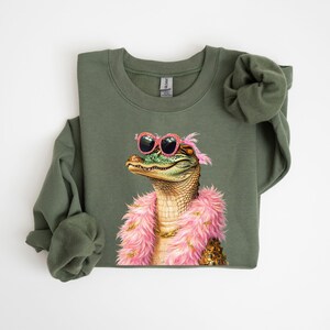 Puede incluir: Sudadera verde oliva con una ilustraci&oacute;n caprichosa de un caim&aacute;n con gafas de sol rosas y una boa rosa. El caim&aacute;n tiene un collar de cadena dorada. Un dise&ntilde;o divertido para una parte superior informal.