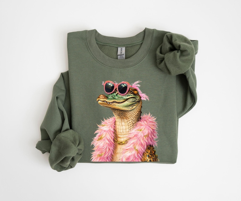 Puede incluir: Sudadera verde oliva con una ilustraci&oacute;n caprichosa de un caim&aacute;n con gafas de sol rosas y una boa rosa. El caim&aacute;n tiene un collar de cadena dorada. Un dise&ntilde;o divertido para una parte superior informal.