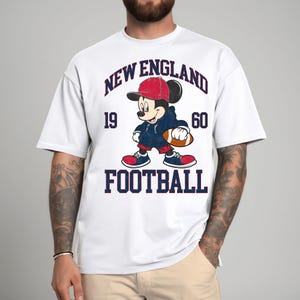 May include: White t-shirt featuring a cartoon Mickey Mouse in a red cap and blue hoodie, holding a football. The shirt has the text "NEW ENGLAND FOOTBALL 1960" in navy blue and red. A fun, sporty design.