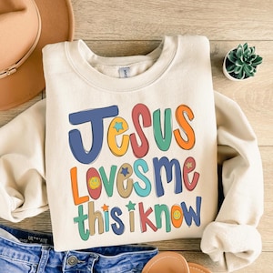 May include: A cream-colored crewneck sweatshirt featuring the colorful text Jesus Loves Me This I Know in a playful, multi-colored font. The design includes small star and smiley face icons interspersed among the letters in shades of blue, orange, red, green, and yellow.