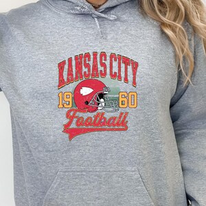 K&ouml;nnte beinhalten: Grauer Kapuzenpullover mit einem Vintage-Grafikdruck eines Footballhelms und dem Text "Kansas City 1960 Football".