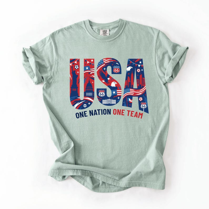 May include: A light sage green t-shirt featuring the large text USA in blue, filled with red and white patriotic graphics including the Statue of Liberty, a bald eagle, and Route 66 signs. Below the text is the phrase ONE NATION ONE TEAM.