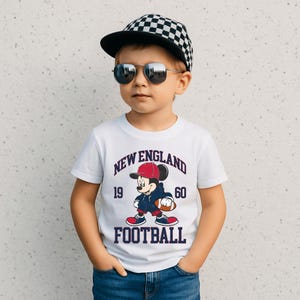 May include: A young child wearing a white t-shirt with a Mickey Mouse graphic in a football theme. The shirt has the words "NEW ENGLAND FOOTBALL 1960". The child is also wearing a checkered baseball cap and sunglasses.