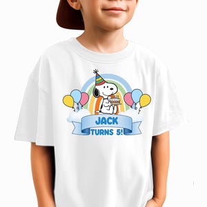 May include: A white t-shirt featuring a graphic of Snoopy wearing a colorful party hat, holding a birthday cake. The design includes a rainbow, balloons, clouds, and a blue banner with the text JACK TURNS 5! printed in bold blue capital letters.