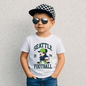 May include: White t-shirt with a graphic of Mickey Mouse in a football uniform, the text "SEATTLE FOOTBALL 19 74". The child is wearing a black and white checkered cap and sunglasses.
