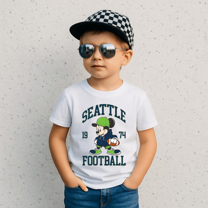 May include: White t-shirt with a graphic of Mickey Mouse in a football uniform, the text "SEATTLE FOOTBALL 19 74". The child is wearing a black and white checkered cap and sunglasses.