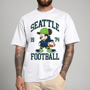 May include: White t-shirt featuring a cartoon Mickey Mouse character in a Seattle Seahawks football uniform. The design includes the words "SEATTLE", "FOOTBALL", and the numbers "19" and "74" in a dark green font. Mickey is wearing a green hat and shoes.