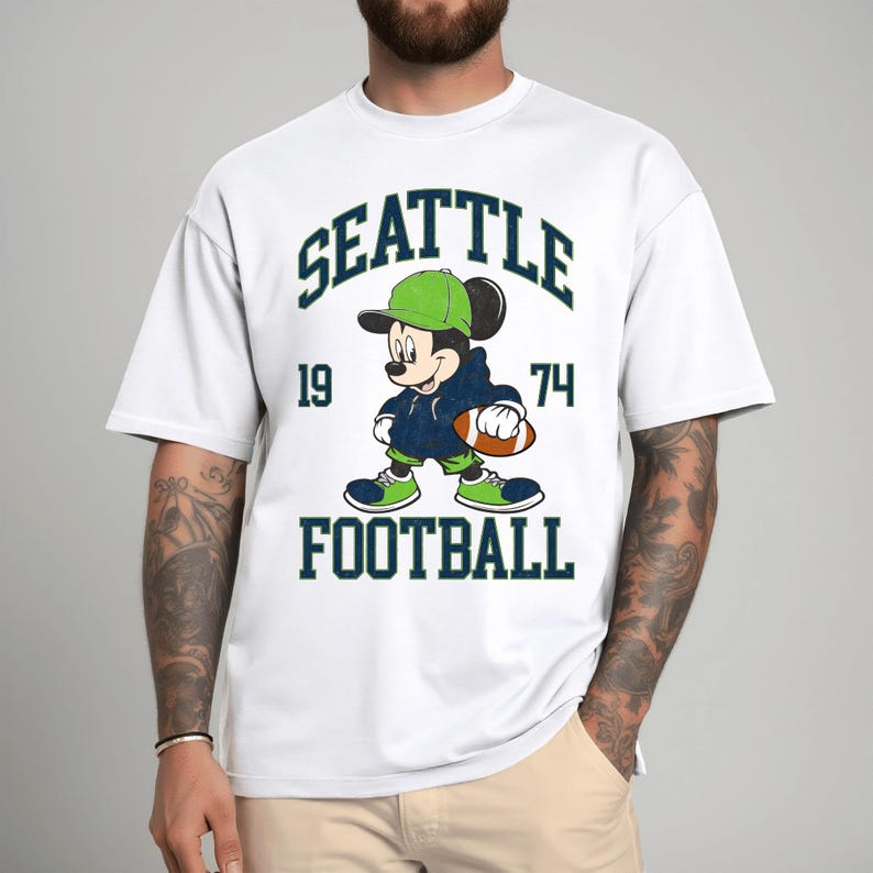 May include: White t-shirt featuring a cartoon Mickey Mouse character in a Seattle Seahawks football uniform. The design includes the words "SEATTLE", "FOOTBALL", and the numbers "19" and "74" in a dark green font. Mickey is wearing a green hat and shoes.
