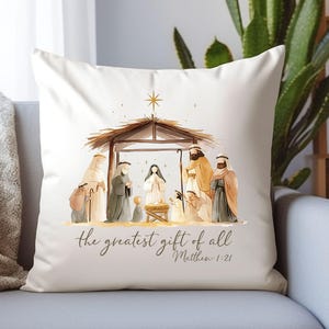 May include: White decorative pillow featuring a watercolor illustration of the Nativity scene. The image includes figures under a wooden structure, with the text "the greatest gift of all Matthew 1:21" in a script font. A star is above the structure.
