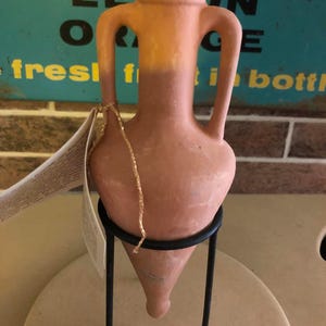 Puede incluir: Una ánfora de color terracota con dos asas, exhibida en un soporte de metal negro. El ánfora tiene un cuerpo redondeado y una base cónica. Una cuerda dorada está unida. El fondo incluye un letrero vintage.