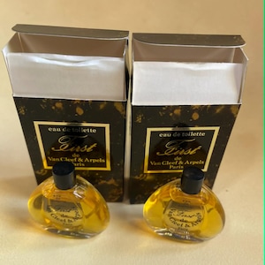 May include: Two miniature perfume bottles with black caps and clear, rounded bodies, filled with a golden liquid. The bottles are in front of their matching boxes, which feature the text "eau de toilette First de Van Cleef & Arpels Paris."