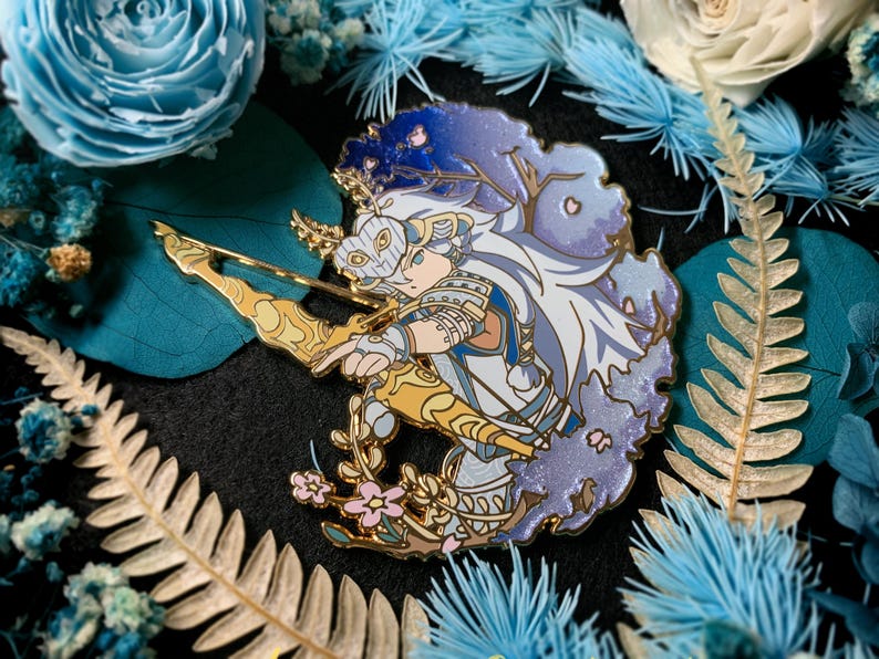 May include: A detailed enamel pin featuring a character with a bow and arrow, set against a backdrop of blue and purple hues. The pin has gold accents and is surrounded by floral and botanical elements. The design is intricate and artistic.