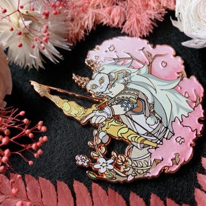 May include: A detailed enamel pin featuring a character with a mask, bow, and arrow, set against a pink cherry blossom backdrop. The pin has gold and white accents, with a floral design at the base. The background includes pink and white flowers.