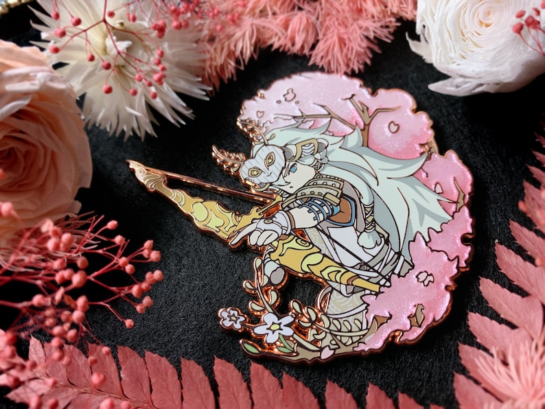 May include: A detailed enamel pin featuring a character with a mask, bow, and arrow, set against a pink cherry blossom backdrop. The pin has gold and white accents, with a floral design at the base. The background includes pink and white flowers.