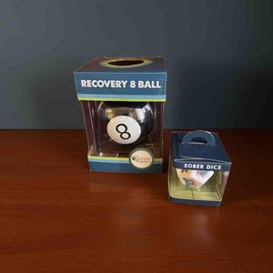 May include: A black and white 8-ball in a clear plastic box labeled "RECOVERY 8 BALL." Beside it is a small box with the text "SOBER DICE." Both are on a wooden surface.