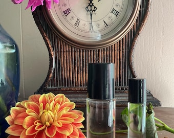 May include: Two clear glass roller bottles with black caps. The larger bottle is in the foreground and the smaller bottle is in the background. The bottles are sitting on a wooden surface. There is a flower in the foreground and a clock in the background.