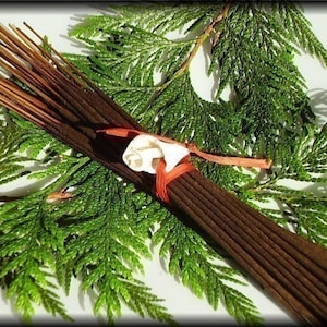 May include: A bundle of brown incense sticks tied together with a red ribbon and a white shell. The incense sticks are surrounded by green cedar branches.